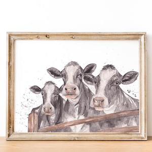 May include: Watercolor painting of three cows with black and white markings, peering over a wooden fence. The artwork is framed in a light-colored wooden frame, set against a white background with subtle paint splatters.