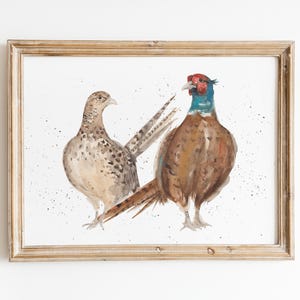 K&ouml;nnte beinhalten: Gerahmtes Aquarellgem&auml;lde mit zwei Fasane. Einer ist braun und beige, der andere hat einen blauen Hals und einen roten Kopf. Das Kunstwerk befindet sich auf einem wei&szlig;en Hintergrund mit kleinen Farbspritzern, in einem hellbraunen Holzrahmen.