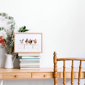 May include: A framed watercolor print featuring five chickens perched on a wooden fence. The artwork is displayed on a wooden desk, alongside a stack of books, a white pitcher with flowers, and a wooden chair.