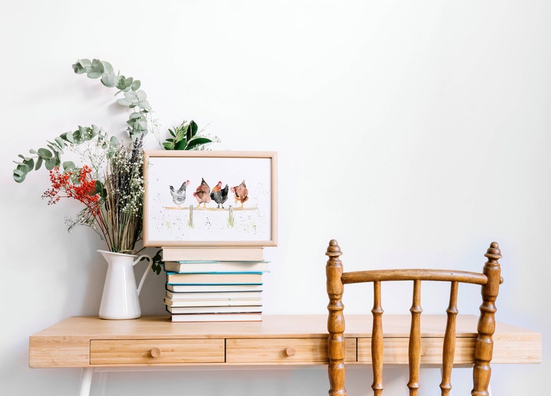 May include: A framed watercolor print featuring five chickens perched on a wooden fence. The artwork is displayed on a wooden desk, alongside a stack of books, a white pitcher with flowers, and a wooden chair.