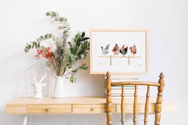 May include: A framed watercolor print featuring five chickens perched on a wooden fence. The chickens are depicted in shades of gray, brown, and black. The artwork is displayed on a light wood desk, alongside a white pitcher with flowers and a decorative deer head.