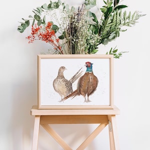 K&ouml;nnte beinhalten: Ein gerahmter Aquarelldruck mit zwei Fasane. Der m&auml;nnliche Fasan hat einen braunen K&ouml;rper, einen blauen Hals und einen roten Kopf. Der weibliche Fasan ist beige mit braunen Flecken. Das Kunstwerk wird auf einem Holzschemel mit einem Blumenstrau&szlig; im Hintergrund ausgestellt.