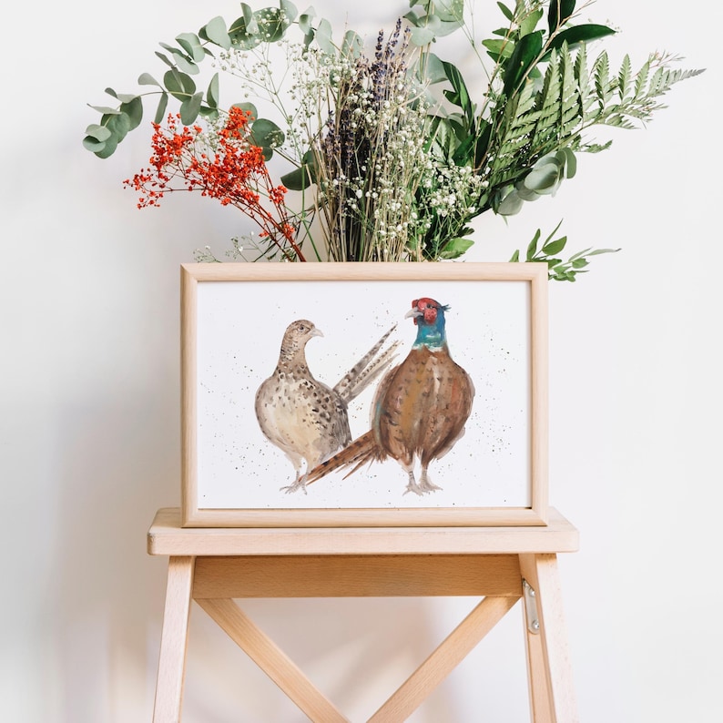 K&ouml;nnte beinhalten: Ein gerahmter Aquarelldruck mit zwei Fasane. Der m&auml;nnliche Fasan hat einen braunen K&ouml;rper, einen blauen Hals und einen roten Kopf. Der weibliche Fasan ist beige mit braunen Flecken. Das Kunstwerk wird auf einem Holzschemel mit einem Blumenstrau&szlig; im Hintergrund ausgestellt.