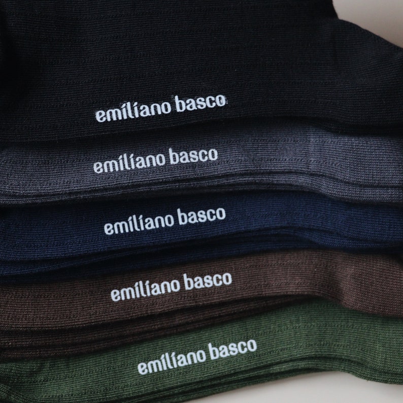 May include: Five pairs of socks in different colors, stacked on top of each other. Each sock has the text "emiliano basco" embroidered on the top in white.