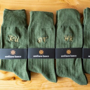 K&ouml;nnte beinhalten: Vier Paar gr&uuml;ne Socken mit gestickten Initialen in einer kursivschrift. Die Socken sind gerippt und haben ein schwarzes Etikett mit dem Text "em&iacute;l&iacute;ano basco" und einem kleinen Logo.