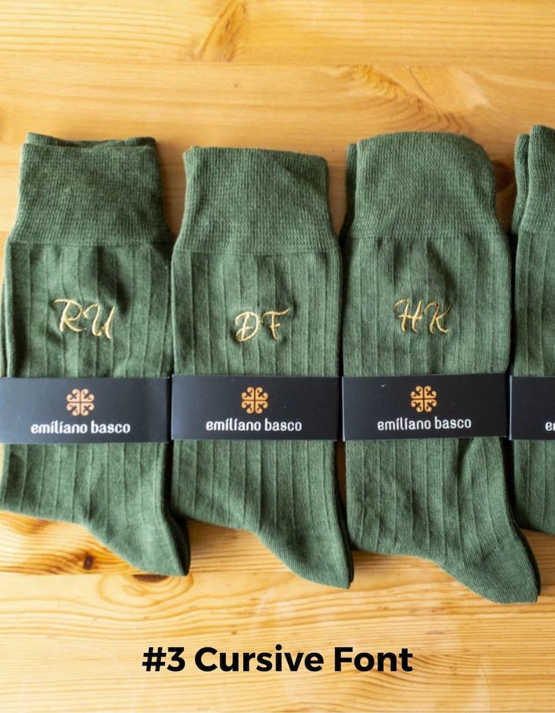 K&ouml;nnte beinhalten: Vier Paar gr&uuml;ne Socken mit gestickten Initialen in einer kursivschrift. Die Socken sind gerippt und haben ein schwarzes Etikett mit dem Text "em&iacute;l&iacute;ano basco" und einem kleinen Logo.