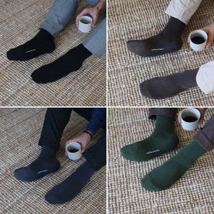 May include: Four pairs of socks in different colors, including black, brown, green, and navy blue. The socks are displayed on a light brown woven surface. Each pair of socks has a small tag with the text "andilana banca" on it.