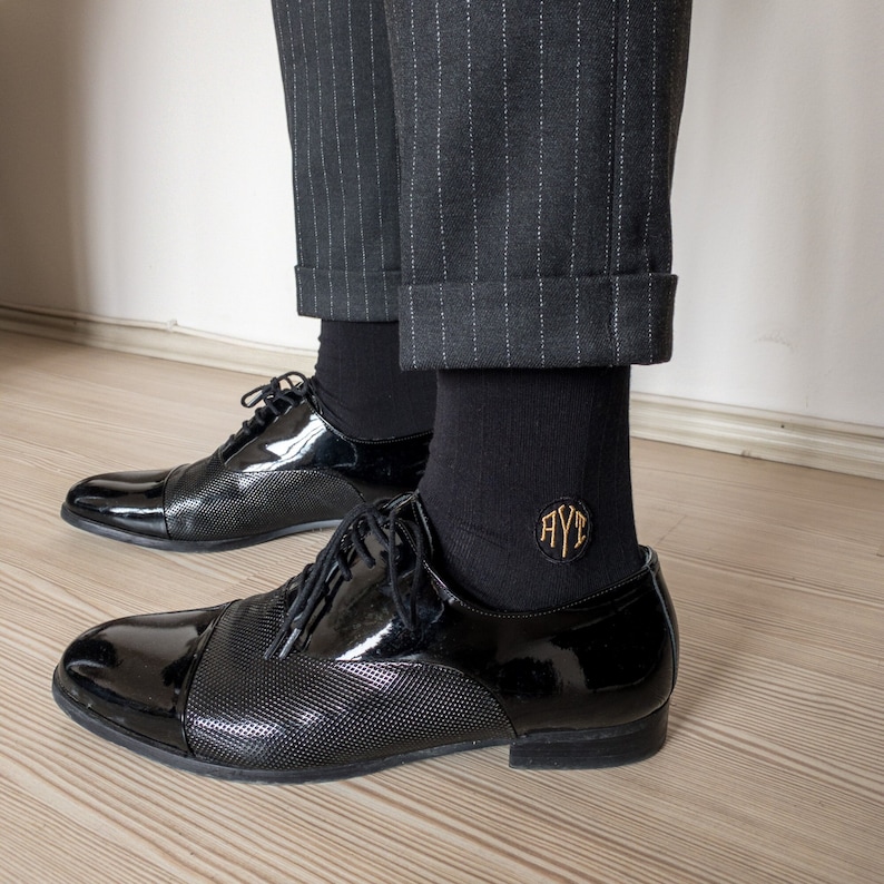 Pu&ograve; includere: Un paio di scarpe da abito in pelle nera con un calzino nero che presenta un cerchio ricamato in oro con le lettere "AYT" all'interno. Le scarpe sono indossate con pantaloni a righe nere.