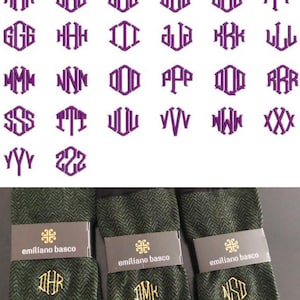 以下が含まれることがあります： 金色の刺繍のイニシャルが入った、緑と黒の模様の靴下3足。靴下は折り畳んで白い表面に置かれています。イニシャルは「DHR」、「DMK」、「NSD」です。