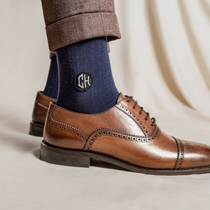 Pu&ograve; includere: Un paio di calzini blu scuro con una toppa monogrammata con le iniziali "CH" in bianco. I calzini sono indossati con scarpe da abito in pelle marrone.