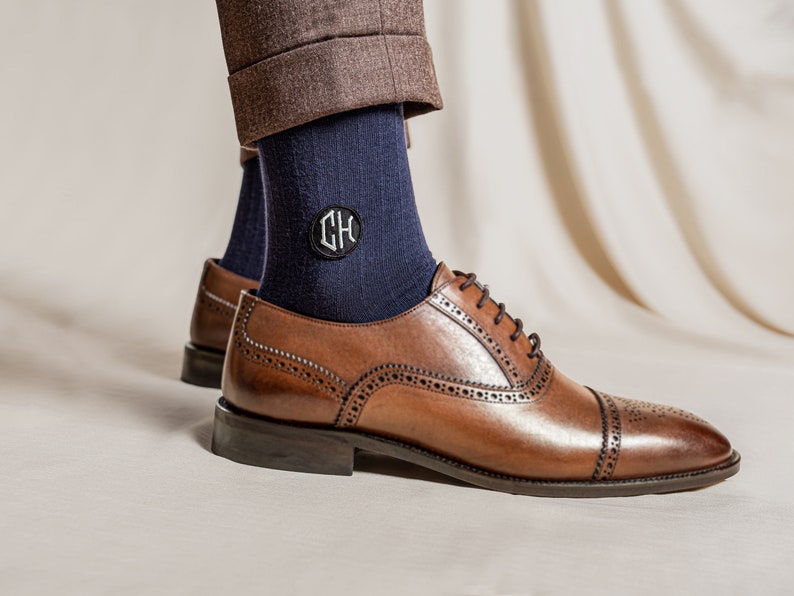 Pu&ograve; includere: Un paio di calzini blu scuro con una toppa monogrammata con le iniziali "CH" in bianco. I calzini sono indossati con scarpe da abito in pelle marrone.