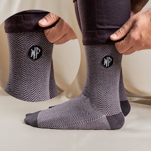 May include: A pair of gray and purple herringbone patterned socks with a dark gray heel and toe. The socks have a black circular patch with the embroidered letters "WF". The socks are being pulled up.