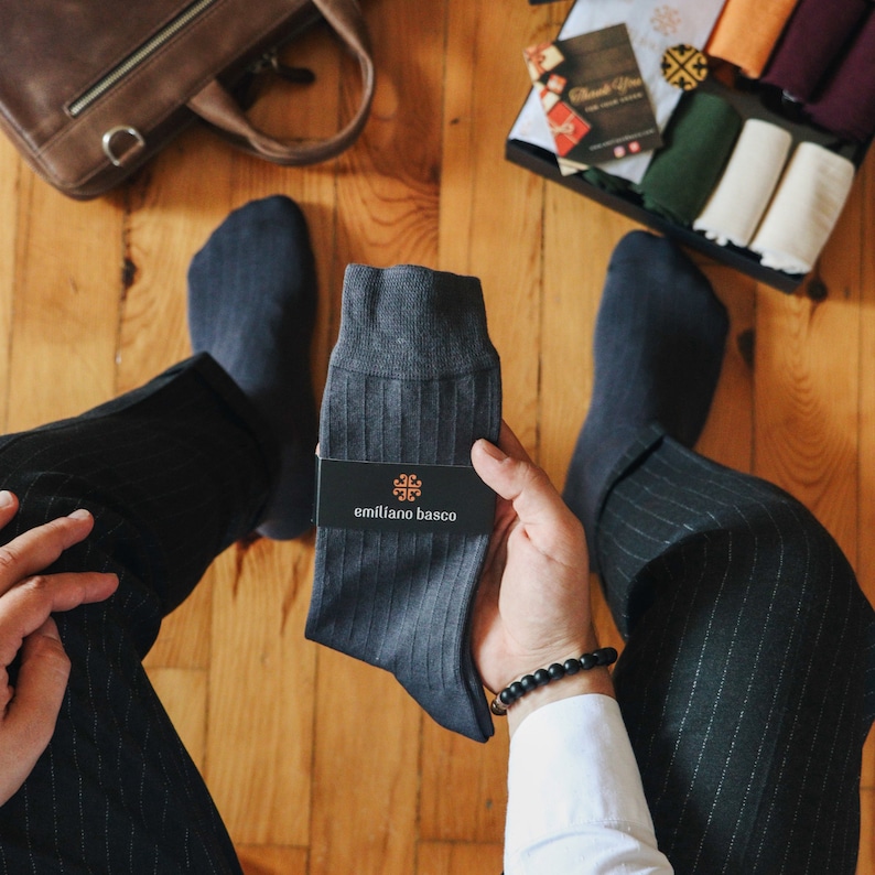 May include: A pair of dark gray ribbed socks with the brand name "emiliano basco" printed on a white tag. The socks are being held by a person wearing black pants and a white shirt.