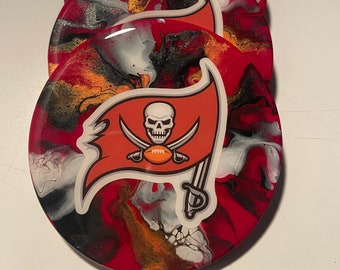 May include: Set of four round coasters with an abstract design in red, black, white and gold. Each coaster features a white Buccaneers logo with a skull and crossbones.