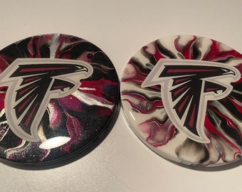 Puede incluir: Dos posavasos redondos con un diseño de remolino negro, blanco y rojo. Los posavasos presentan un logotipo de los Atlanta Falcons plateado y rojo en el centro.