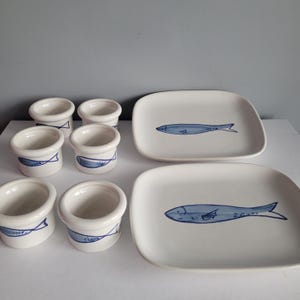 May include: A collection of white ceramic tableware with a nautical theme. The set includes small, cylindrical cups and square plates, each decorated with a blue fish illustration. The cups feature a blue band design. Suitable for serving appetizers or small portions.
