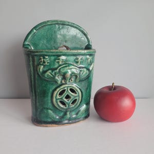 May include: A green ceramic wall pocket with a semi-circular top, featuring a decorative face and a circular design with cutouts. A red apple with a stem sits beside it.