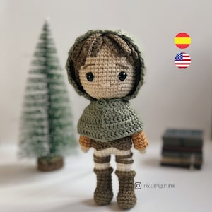 Puede incluir: Una muñeca de crochet con pelo marrón, que lleva una capa verde con capucha, pantalones marrones y botas naranja y marrones. La muñeca tiene un botón en su capa y está de pie sobre una superficie blanca.