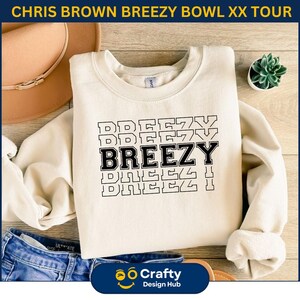 May include: Cream-colored sweatshirt with the word "BREEZY" in a bold, black, stacked design. The sweatshirt also features the text "CHRIS BROWN BREEZY BOWL XX TOUR" at the top. The design is printed on a soft, casual garment.