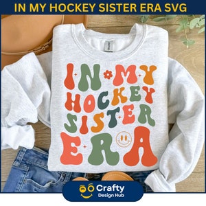 May include: A gray sweatshirt with the text "IN MY HOCKEY SISTER ERA" in colorful, retro-style lettering. The letters are outlined in white and have a slightly raised, 3D effect.