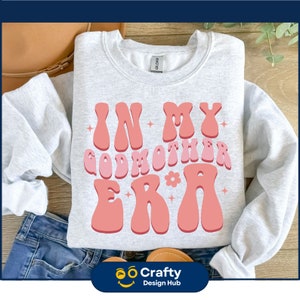 Op de afbeelding: Een lichtgrijze sweatshirt met de tekst "IN MY GODMOTHER ERA" in een retro lettertype. De tekst is in roze en perziktinten, met ster- en bloemaccenten. De sweatshirt is opgevouwen op een oppervlak met een jeans en een handtas.