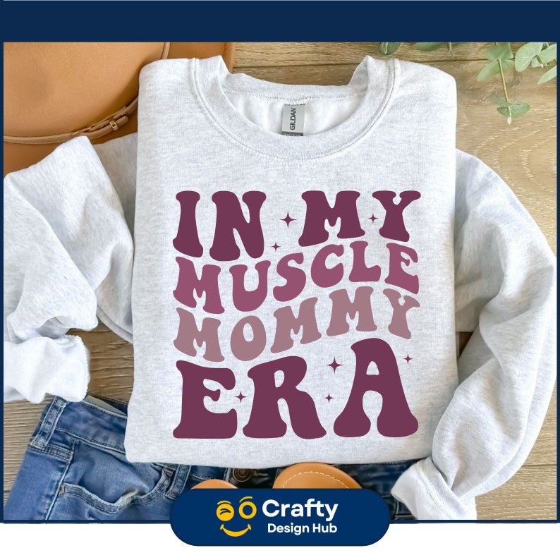 May include: A light gray sweatshirt with the text "IN MY MUSCLE MOMMY ERA" in shades of purple. The sweatshirt is folded, with the sleeves visible. The Crafty Design Hub logo is at the bottom.