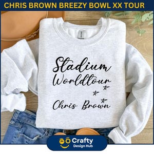 May include: A light gray sweatshirt with the text "Stadium Worldtour Chris Brown" in black script. The sweatshirt has a crew neck and long sleeves. The image also includes a pair of blue jeans and a brown purse.