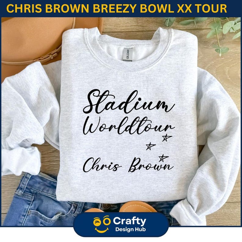 May include: A light gray sweatshirt with the text "Stadium Worldtour Chris Brown" in black script. The sweatshirt has a crew neck and long sleeves. The image also includes a pair of blue jeans and a brown purse.