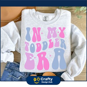 以下が含まれることがあります： 星形のピンクとブルーのレトロな文字で「In My Toddler Era」と書かれたグレーのトレーナーです。