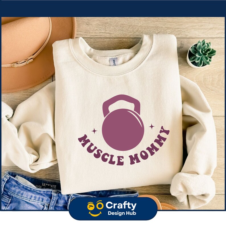 May include: Cream-colored sweatshirt with a purple kettlebell graphic and the words "Muscle Mommy". The design includes two small star accents. The sweatshirt is displayed on a wooden surface with a hat and jeans.