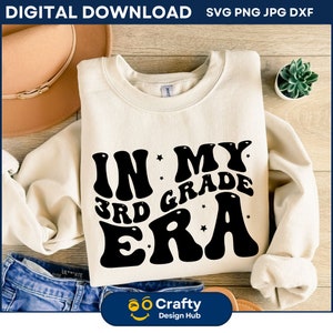 Könnte beinhalten: Ein weißes Sweatshirt mit schwarzem Text, der "In My 3rd Grade Era" mit Sternen um den Text herum lautet. Das Sweatshirt liegt auf einer Holzoberfläche mit einer Jeans, einem Hut und einer Topfpflanze.