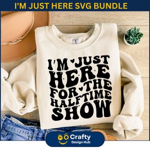 May include: A white sweatshirt with the text "I'm Just Here For The Halftime Show" in black retro-style lettering. The text is arranged in a circular pattern with a heart symbol between the words "I'm" and "Just" and between the words "For" and "The".