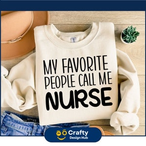 May include: A white sweatshirt with the text "My Favourite People Call Me Nurse" printed in black. The sweatshirt is laid out on a wooden surface with a brown hat and a potted plant.