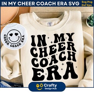 May include: A white sweatshirt with a black graphic design that says "IN MY CHEER COACH ERA" in a retro font. The graphic is surrounded by small stars. The sweatshirt is on a wooden surface.