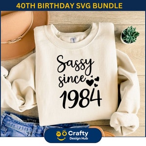 May include: A white sweatshirt with the text "Sassy since 1984" in black lettering. The text is in a cursive font and includes small hearts around the year.