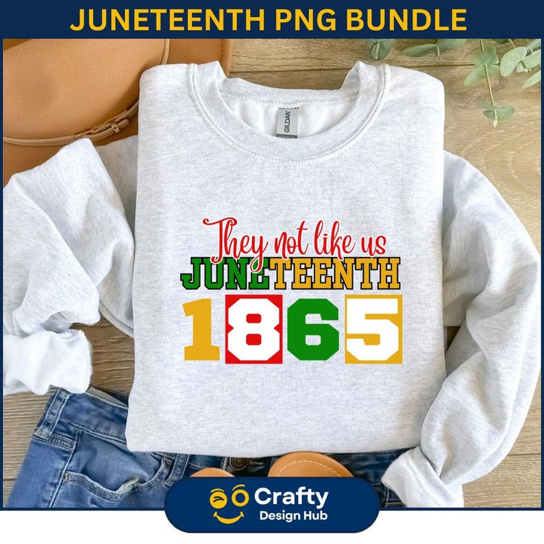 Pu&ograve; includere: Una felpa grigio chiaro con la scritta "They not like us Juneteenth 1865" in rosso, verde, giallo e bianco. La felpa &egrave; piegata su una superficie di legno con un paio di jeans.
