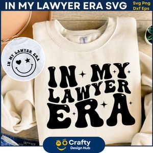 May include: A white sweatshirt with the text "IN MY LAWYER ERA" in black, stylized lettering. The text is in a retro, groovy font with stars and hearts around it. The sweatshirt is laying flat on a wooden surface.