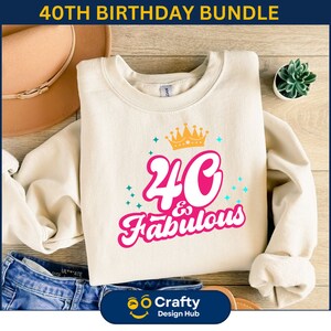 May include: A white sweatshirt with a pink graphic design that says "40 & Fabulous" with a gold crown above the text. The sweatshirt is on a wooden surface with a hat and a potted plant.