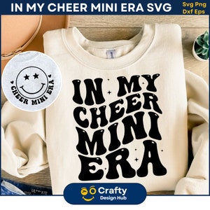 May include: A white sweatshirt with a black graphic design that says "In My Cheer Mini Era" in a retro font. The graphic is surrounded by small stars. The sweatshirt is folded and laid on a wooden surface.