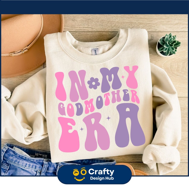 Op de afbeelding: Cr&egrave;mekleurige sweatshirt met de tekst "IN MY GODMOTHER ERA" in roze en paarse retro lettertype. Het ontwerp bevat bloemen- en steraccenten. De sweatshirt ligt op een houten oppervlak met een hoed en jeans.