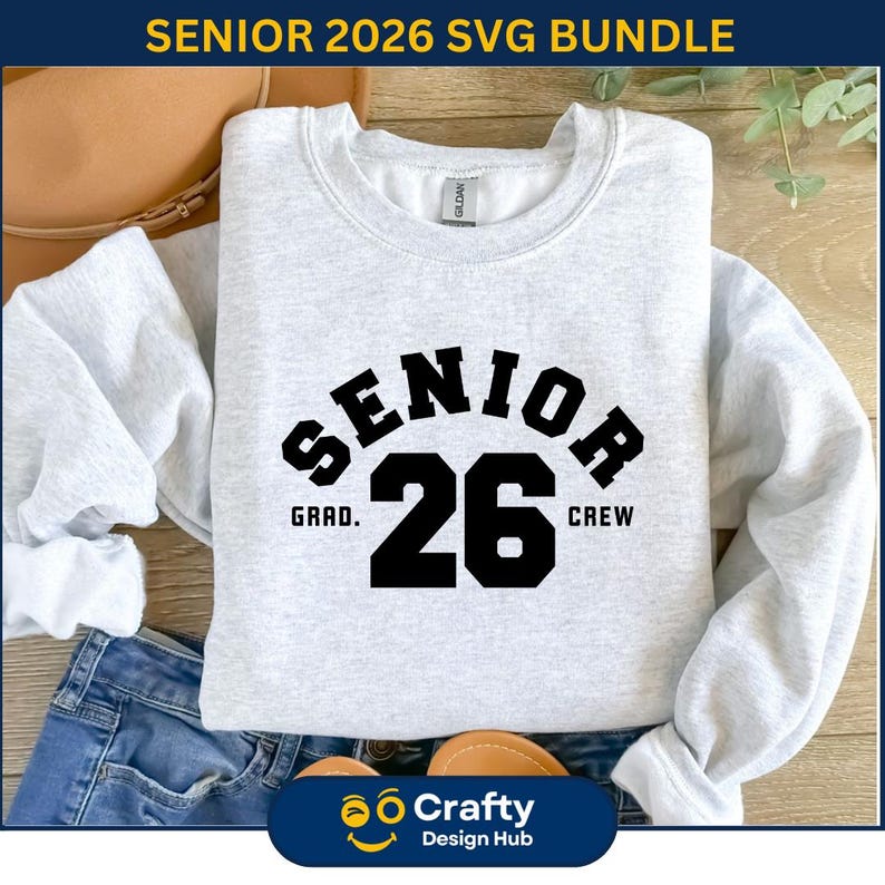 May include: A light gray sweatshirt with the text "SENIOR 26" in black block letters. The word "SENIOR" arches above the number "26", with "GRAD." and "CREW" on either side. The sweatshirt is folded on top of blue jeans.