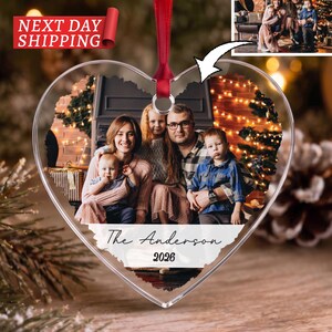 May include: A heart-shaped clear acrylic Christmas ornament featuring a printed family photograph of four people in a festive setting. The ornament includes a red ribbon for hanging and white text at the bottom that reads The Anderson 2026.