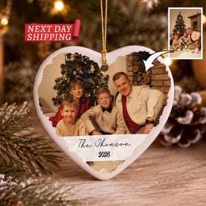 May include: A heart-shaped ceramic ornament featuring a printed photograph of a family of four in front of a Christmas tree. The ornament includes a gold hanging string and the text The Thomson 2026 printed in black script and serif font.