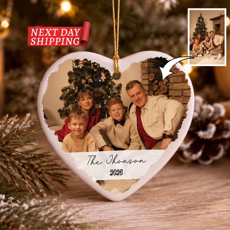 May include: A heart-shaped ceramic ornament featuring a printed photograph of a family of four in front of a Christmas tree. The ornament includes a gold hanging string and the text The Thomson 2026 printed in black script and serif font.