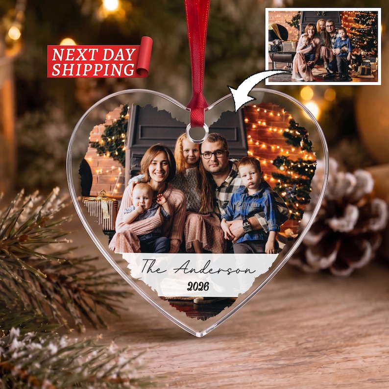 May include: A heart-shaped clear acrylic Christmas ornament featuring a printed family photograph of four people in a festive setting. The ornament includes a red ribbon for hanging and white text at the bottom that reads The Anderson 2026.