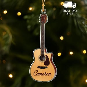 May include: Acoustic guitar ornament with a natural wood finish, brown trim, and the name "Cameron" in a cursive font. The ornament is hanging from a gold string. The background is a blurred green with bokeh lights.