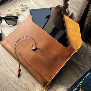 May include: A brown leather envelope-style case with a black tablet computer inside. The case has a flap with a brown leather strap and a gold metal button. The case is on a wooden surface.
