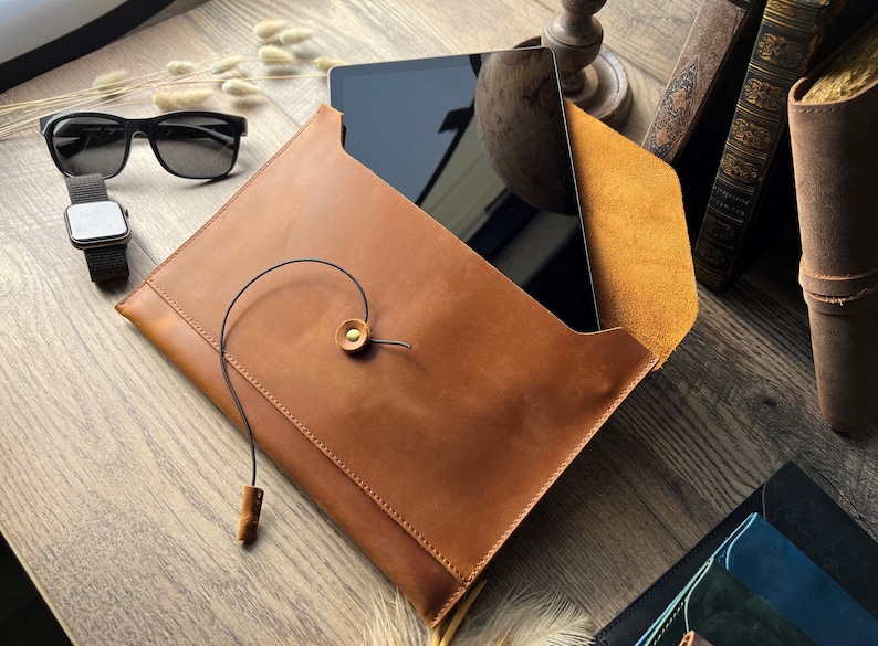 May include: A brown leather envelope-style case with a black tablet computer inside. The case has a flap with a brown leather strap and a gold metal button. The case is on a wooden surface.