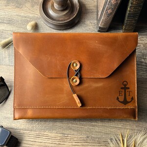 May include: A brown leather laptop sleeve with a flap closure and a black elastic strap with two brass buckles. The sleeve has an engraved anchor with the initials "ET".