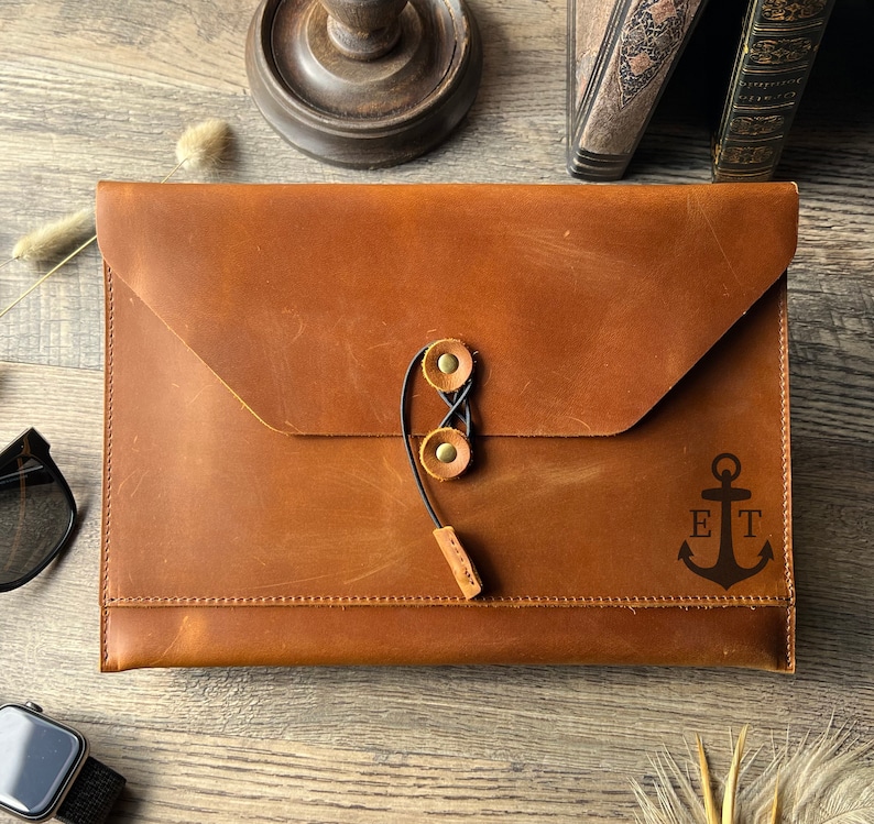 May include: A brown leather laptop sleeve with a flap closure and a black elastic strap with two brass buckles. The sleeve has an engraved anchor with the initials "ET".